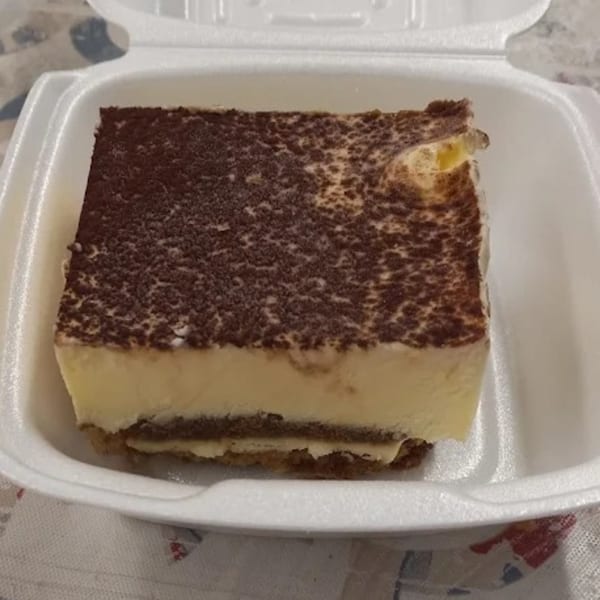 A slice of tiramisu with cocoa powder dusting on top, served in a white takeout container.